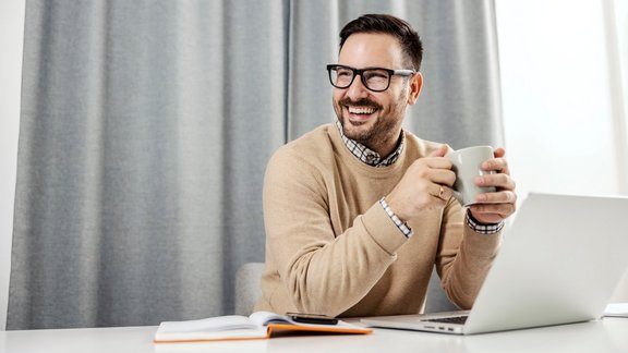 Ein glücklicher Professional Scrum Master sitzt in seinem Homeoffice, hält eine Tasse Kaffee hoch und macht eine Pause.