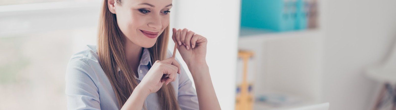 Textverarbeitung mit Word Porträt einer charmanten, selbstbewussten, netten, lächelnden Frau mit Brille auf dem Kopf, die einen Stift in den Händen hält und auf den Bildschirm des Computers schaut, während sie am Schreibtisch an ihrem Arbeitsplatz sitzt.