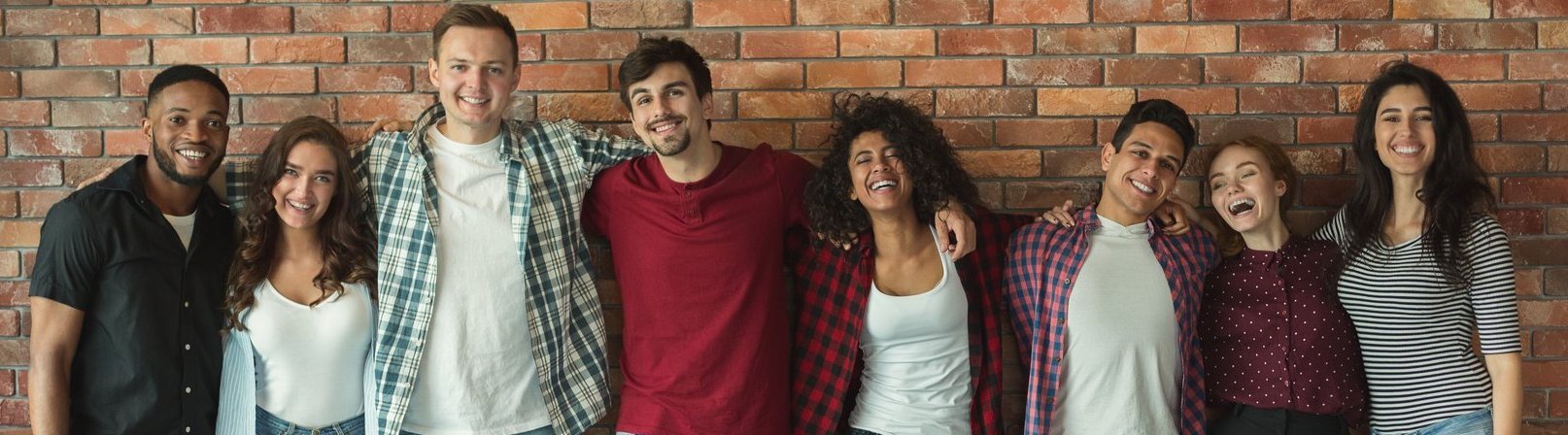 Maßnahmekombination Einzelcoaching Fröhliche multikulturelle Freunde, die lachen und sich umarmen, während sie für ein Gruppenporträt posieren. Sie stehen versammelt vor einer roten Backsteinmauer.