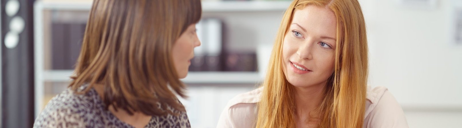 Vorbereitung auf eine Umschulung Zwei Geschäftsfrauen in einer ernsten Diskussion sitzen gemeinsam an einem Schreibtisch und schauen auf Unterlagen, mit Fokus auf eine attraktive junge Frau. Die beiden bereiten sich im Kurs 'Vorbereitung auf eine Umschulung' auf den Wiedereinstieg in den Lernalltag vor.
