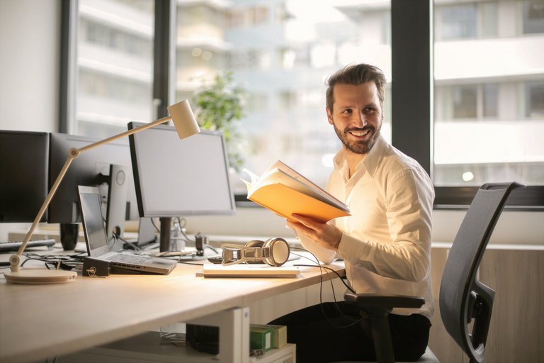 Mann im Büro sitzt vor einer Fensterfront an einem Schreibtisch mit Computer. Er hat sich umgedreht, ein Buch in der Hand und lächelt.