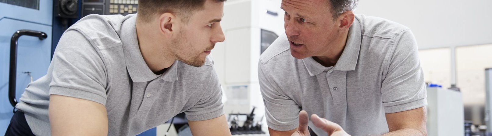 Assistierte Ausbildung Intensives Gespräch zwischen Schichtleiter und Arbeiter. Beide sind nach vorne auf einen Arbeitstisch mit Werkstücken aus Metall gelehnt.