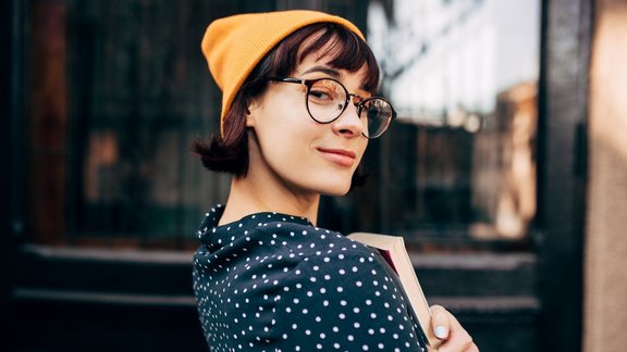 Porträt einer jungen Studentin mit gelbem Hut, transparenten Brillen und einem grünen Hemd mit weißen Punkten, die Bücher in den Händen hält und vor der Universität steht.