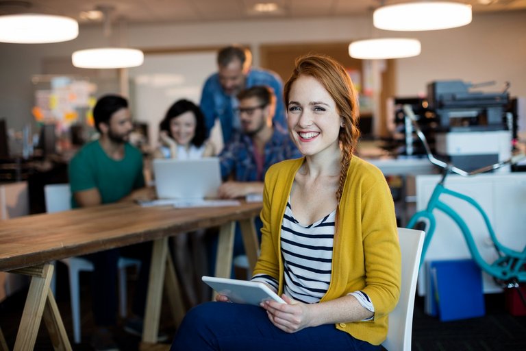 Eine hübsche Frau benutzt ein digitales Tablet und lächelt in die Kamera, während im Hintergrund das Team arbeitet und unscharf zu sehen ist.
