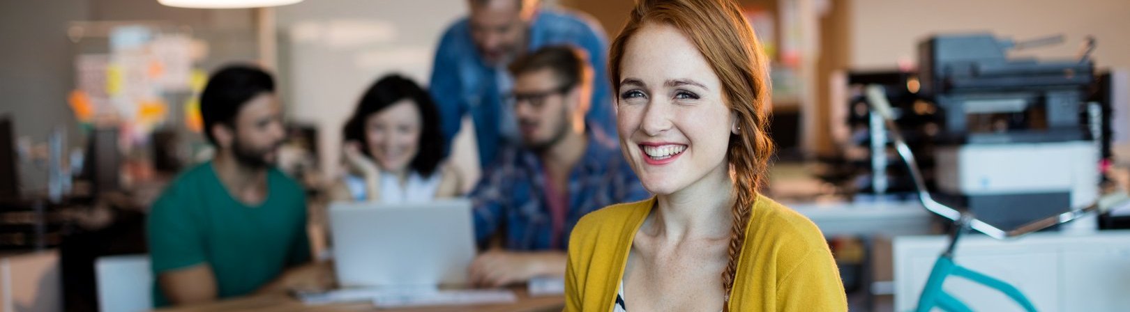 Eine hübsche Frau benutzt ein digitales Tablet und lächelt in die Kamera, während im Hintergrund das Team arbeitet und unscharf zu sehen ist.