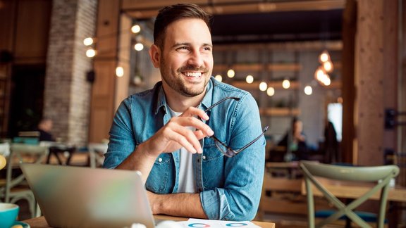 Lächelnder Mann sitzt im Café vor einem Laptop und schaut aus dem Fenster – Teilnehmer des Gründercoachings bei der Arbeit an seinem Businessplan.