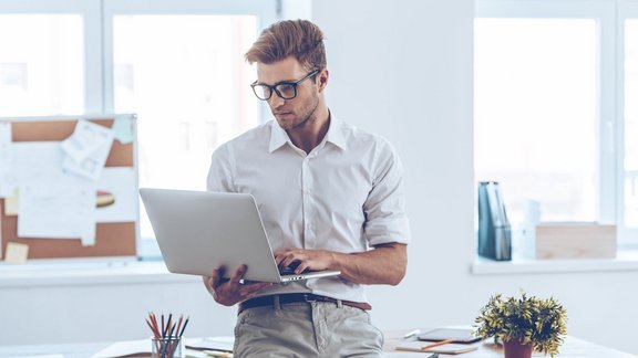Gepflegter junger Mann mit Brille, der seinen Laptop benutzt, während er sich am Tisch im Büro lehnt.