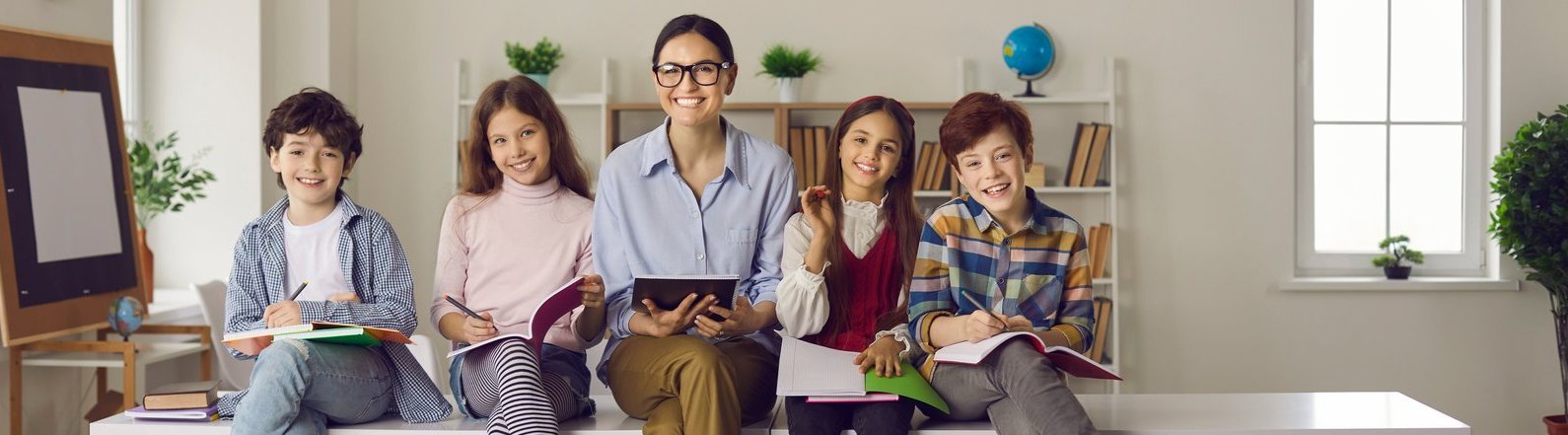 Schulbegleiter mit pädagogischer Zusatzqualifikation Eine fröhliche junge Lehrerin und verschiedene Kinder schauen in die Kamera, während sie auf dem Klassenzimmertisch mit Schreibutensilien und Büromaterial sitzen.