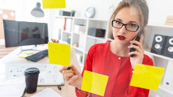 Junge Frau mit Brille und rotem Shirt arbeitet im Büro mit Post-its. Foto mit Tiefenschärfe.