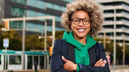 Gut gelaunte Frau mit Brille und grünem Hoodie steht vor unscharfer Stadtkulisse im Hintergrund.