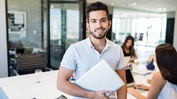 Lächelnder junger hispanischer Geschäftsmann sitzt mit einem Laptop an einem Schreibtisch im Büro – Teilnehmer des Kurses 'Grundlagen der Volks- und Betriebswirtschaftslehre für Industriekaufleute', um wirtschaftliche Schlüsselkonzepte zu erlernen.