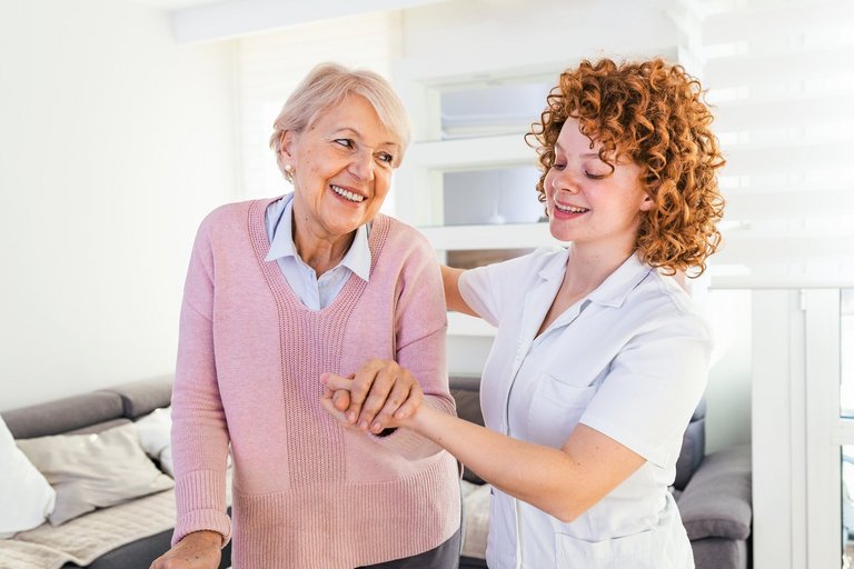 Lächelnde Pflegekraft hilft einer älteren Dame, im Pflegeheim herumzulaufen. Porträt einer glücklichen weiblichen Pflegekraft und einer Seniorin, die gemeinsam zu Hause spazieren gehen.