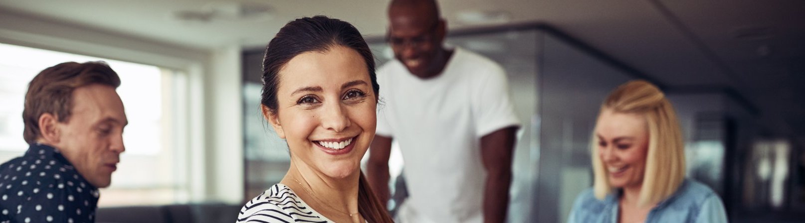 Kaufmännisches Englisch – A2 Eine junge Frau schaut über ihre Schulter und lächelt, während sie mit einer Gruppe von anderen Teilnehmenden an einem Tisch sitzt während des Kurses 'Kaufmännisches Englisch A2'. Es gibt eine Gruppenübung.
