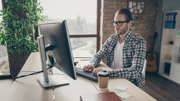 Foto eines Mannes, der auf den Computermonitor am Tisch schaut, und im Homeoffice arbeitet. Er trägt ein lässiges Hemd und eine Brille.