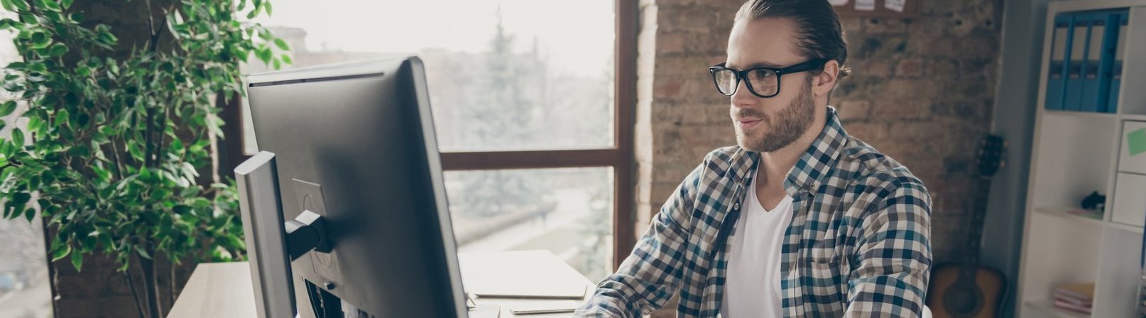 Psychologischer Berater – Schwerpunkt Kinder- und Jugendberatung Foto eines Mannes, der auf den Computermonitor am Tisch schaut, und im Homeoffice arbeitet. Er trägt ein lässiges Hemd und eine Brille.