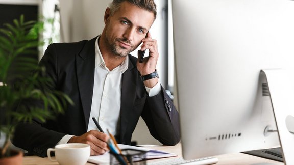 Ein erwachsener Geschäftsmann in seinen 30ern, der einen Anzug trägt, am Handy spricht und gleichzeitig im Büro am Computer arbeitet.