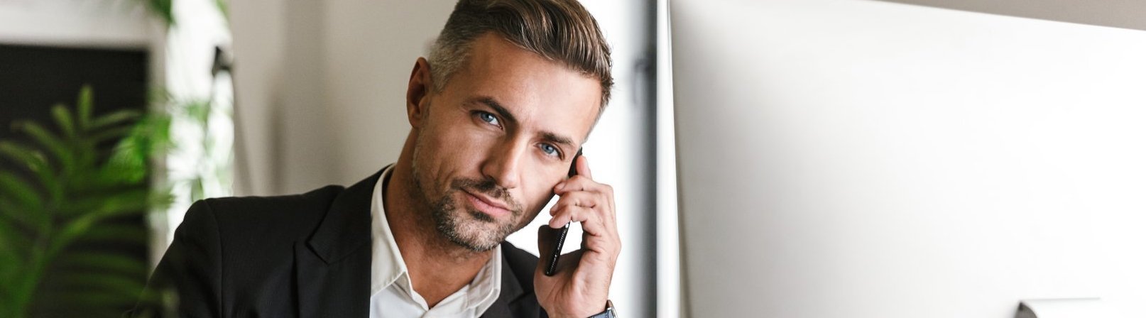 Personalreferent Ein erwachsener Geschäftsmann in seinen 30ern, der einen Anzug trägt, am Handy spricht und gleichzeitig im Büro am Computer arbeitet.