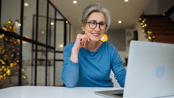 Glückliche, reife Frau mittleren Alters, stützt ihren Kopf auf einer Hand ab und blickt auf den Bildschirm ihres Laptops.