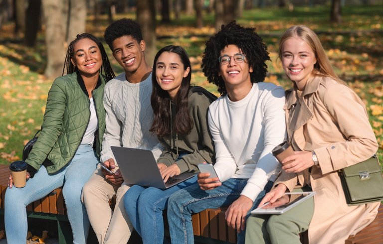 Fröhliche internationale Teenager-Freunde sitzen auf einer Bank mit ihren Geräten und verbringen Zeit zusammen im Park.