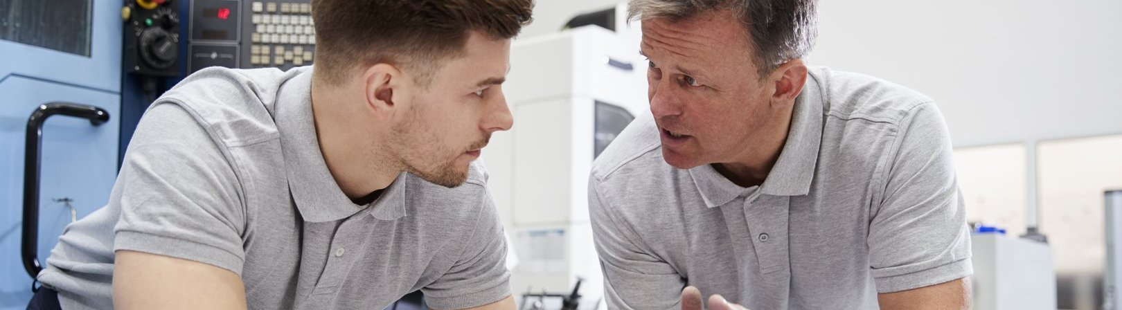 Ein Ingenieur zeigt einem Lehrling, wie man CAD-Zeichnungen misst. Beide sind nach vorne auf den Tisch gelehnt und unterhalten sich angeregt.