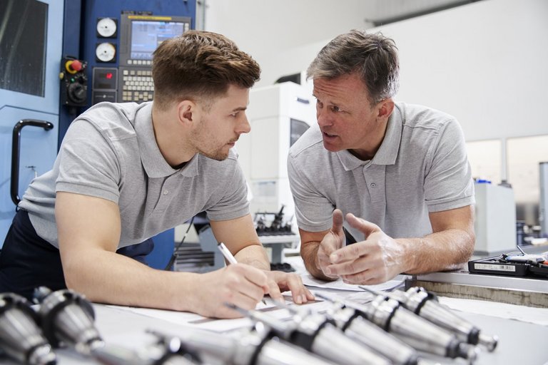 Zwei Techniker in einer Werkstatt lehnen über einen Tisch und diskutieren.