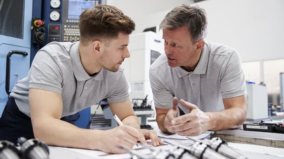 Zwei Techniker in einer Werkstatt lehnen über einen Tisch und diskutieren.