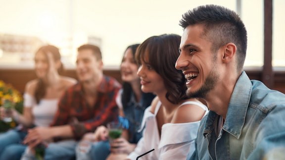 Eine Gruppe junger, lächelnder Menschen trinkt Cocktails und entspannt sich auf einer Dachterrasse bei Sonnenuntergang.
