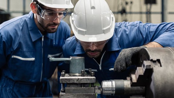 2 Ingenieure in Arbeitskleidung und Sicherheitsausrüstung, der an einer Drehmaschine im Werk mit Metall arbeitet.