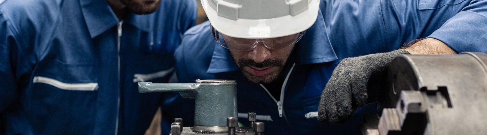 Umschulung Fachkraft Metalltechnik | Fachrichtung Zerspanungstechnik 2 Ingenieure in Arbeitskleidung und Sicherheitsausrüstung, der an einer Drehmaschine im Werk mit Metall arbeitet.
